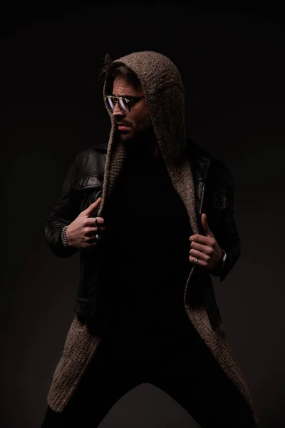 sexy young man with wool cardigan and glasses arranging leather jacket and looking to side while confidently posing on grey background
