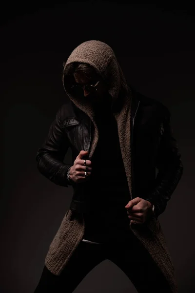 mysterious man with hoodie and leather jacket looking down, pulling jacket and posing in a cool way on grey background