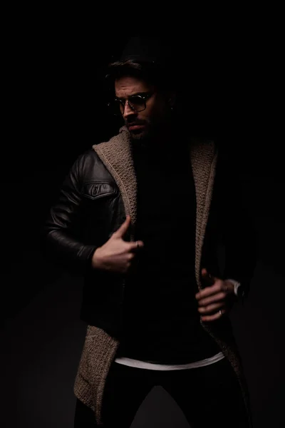 mysterious cool guy looking away and adjusting leather jacket and wool cardigan on grey background
