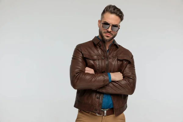 portrait of a attractive casual man with tough look is crossing his arms, standing, wearing sunglasses against gray studio background