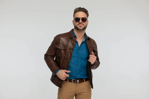 portrait of a attractive casual man with style is fixing his jacket, standing, wearing sunglasses against gray studio background