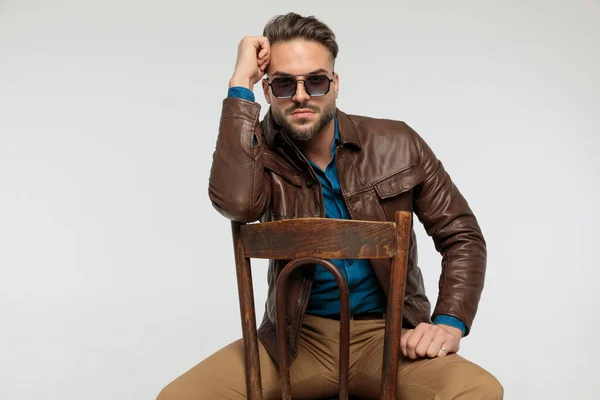 portrait of a attractive casual man resting his arms and touching his forehead, sitting on a chair, wearing sunglasses against gray studio background