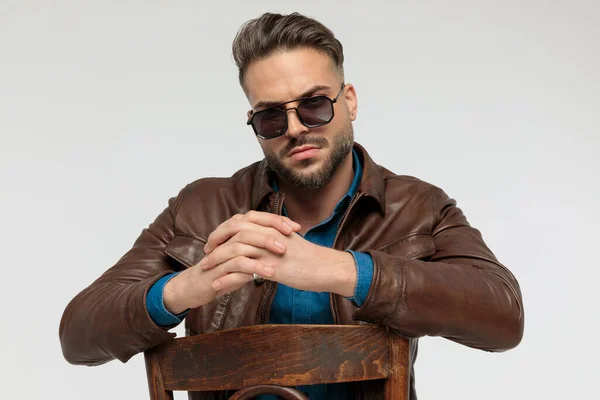 portrait of a attractive casual man resting his arms together with tough attitude, sitting on a chair, wearing sunglasses against gray studio background