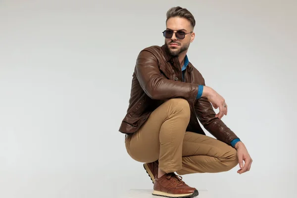 portrait of a attractive casual man resting arms, squatting and looking away, standing, wearing sunglasses against gray studio background