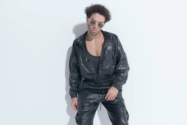 fashion cool guy with curly hair wearing leather suit and sunglasses laying on a wall and posing on grey background