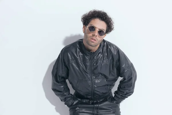 turkish man with curly hair in leather suit holding hands in pockets and laying on a grey wall