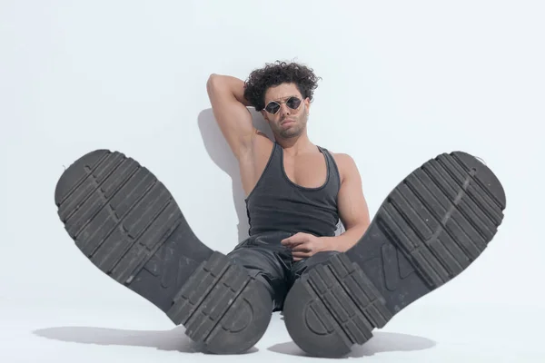 confident muscle guy with curly hair holding hand behind neck, showing boots and sitting on grey background