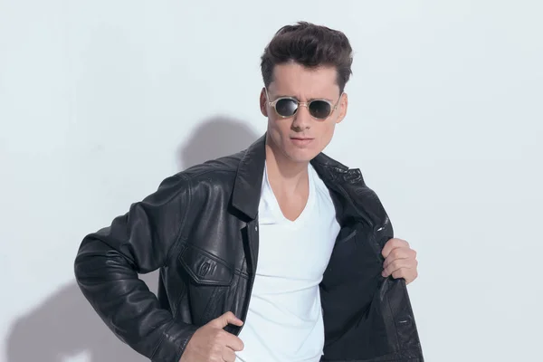 Portrait of  attractive casual man with macho style is pulling his leather jacket, standing, wearing sunglasses in a fashion pose