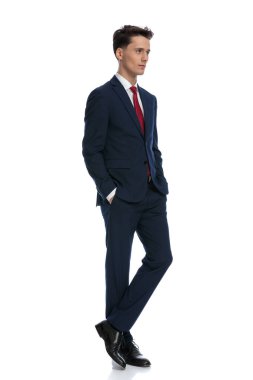 elegant businessman with hands in pockets crossing his legs, wearing a suit and tie against white background
