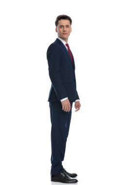young businessman turning to the side and looking at camera, wearing a suit and tie against white background