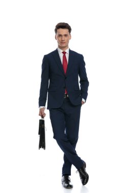 handsome businessman crossing legs, holding a briefcase with hand in pocket, wearing a suit and tie against white background