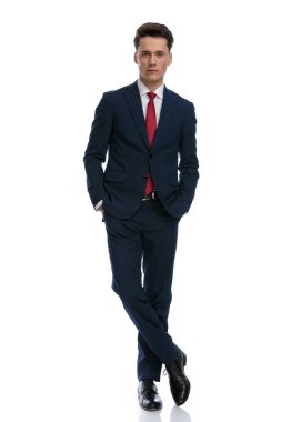 handsome businessman holding hands in pockets and crossing legs, wearing a suit and tie against white background