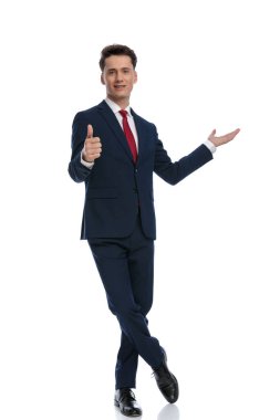 young businessman pointing to side, giving thumbs up with crossed feet, wearing a suit and tie against white background