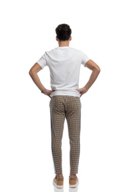 behind view of casual man with plaid pants and white t-shirt posing with hands on hips in front of white background in studio