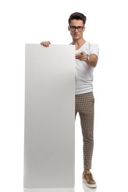 full body picture of young man with glasses showing empty board and pointing finger on white background