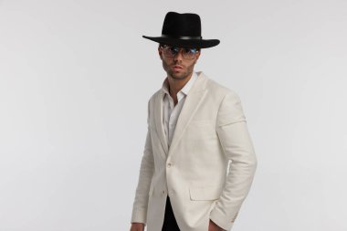 Portrait of attractive smart casual man with nice style is posing, standing, wearing eyeglasses and black hat against gray background