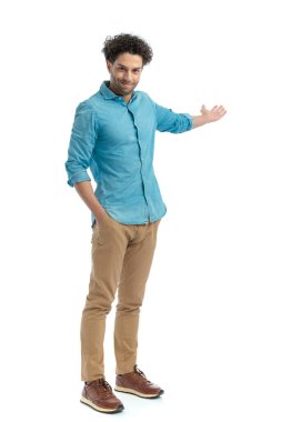 handsome greek guy with hands in chino pants showing to side and smiling while standing with one hand in pocket on white background