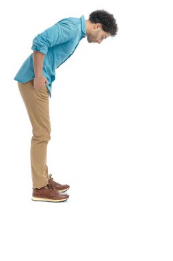 side view of curly hair man with hands in pockets looking down trying to see something small on the floor in front of white background