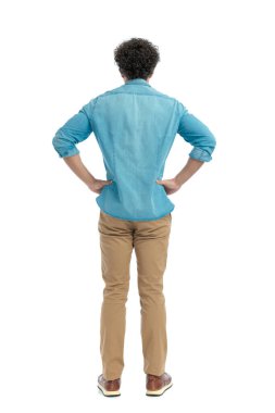 back view of casual young man holding hands on hips and standing in front of white background in studio