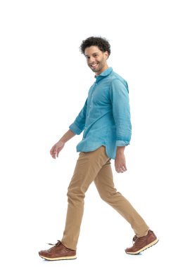 side view of proud casual man with curly hair smiling and walking in front of white background in studio