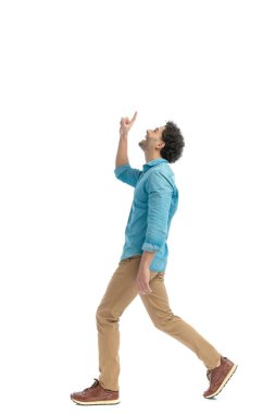 side view of thankful young man pointing finger to the sky, walking and smiling in front of white background