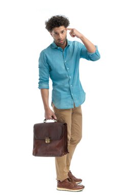upset young man holding suitcase and pointing finger to temple while standing on white background in studio