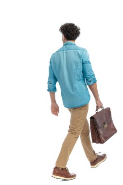 rear view of turkish man with curly hair holding suitcase and walking in front of white background in studio