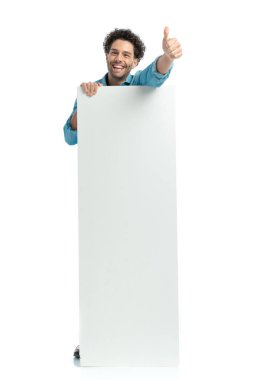 happy arab man covering his body with empty board and laughing while making thumbs up gesture on white background