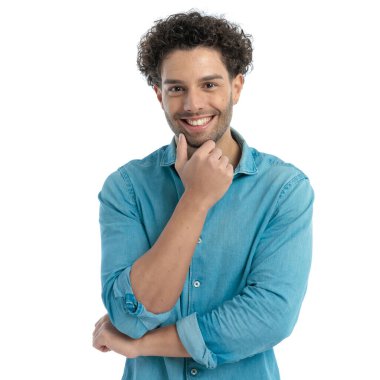 happy young man with curly hair holding hand to chin and thinking in front of white background in studio
