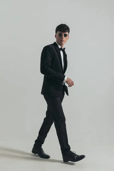 Full body picture of attractive businessman with badass walk, standing, wearing a black tuxedo and sunglasses, in a fashion pose
