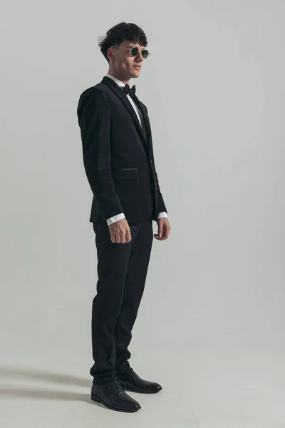Full body picture of young businessman looking away and posing, standing, wearing a black tuxedo and sunglasses, in a fashion pose