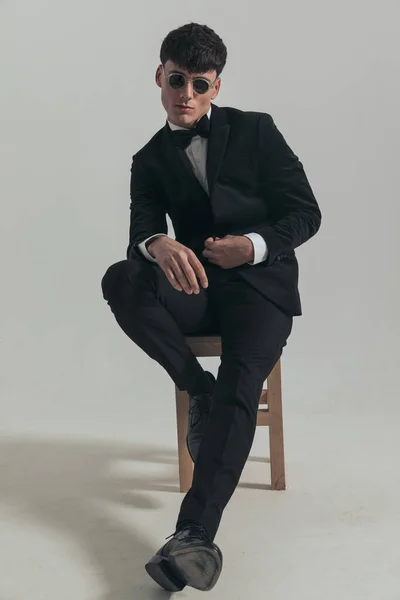 Full body picture of attractive businessman closing his jacket slowly, sitting on a wooden chair, wearing a black tuxedo and sunglasses, in a fashion pose