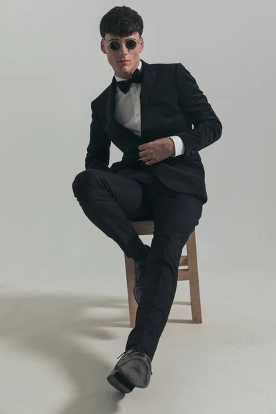 Full body picture of young businessman opening his jacket and dominating the frame, sitting on a wooden chair, wearing a black tuxedo and sunglasses, in a fashion pose