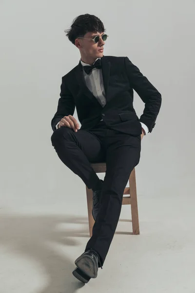 Full body picture of handsome businessman looking over his shoulder, sitting on a wooden chair, wearing a black tuxedo and sunglasses, in a fashion pose