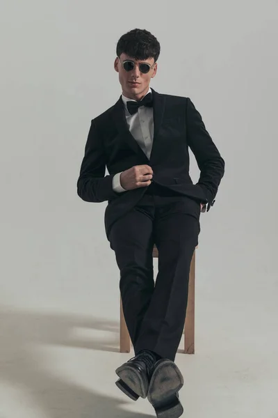 Full body picture of handsome businessman relaxing while closing his jacket, sitting on a wooden chair, wearing a black tuxedo and sunglasses, in a fashion pose