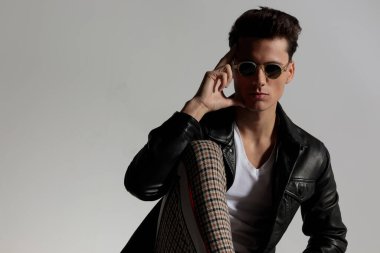 thoughtful cool guy with leather jacket holding fingers to temple and thinking while posing with knee up in front of grey background