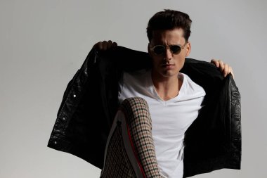 stylish young man pulling and adjusting black leather jacket while posing in front of grey background in studio
