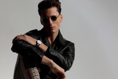 happy cool guy with plaid pants and leather jacket posing with elbow on knee on grey background