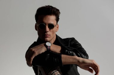 portrait of cool stylish man in leather jacket holding elbow on knee and posing in front of grey background