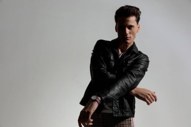 handsome casual man in black leather jacket holding arms in fashion arms and posing in front of grey background in studio