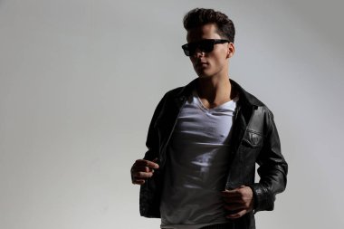 sexy cool guy with glasses looking away and adjusting lack leather jacket in front of grey background in studio