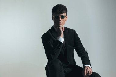 attractive young man in elegant suit holding elbow on knee and posing in front of grey background in studio