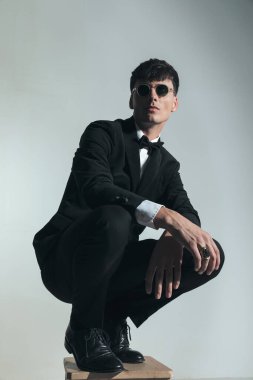 cool elegant best man with glasses wearing black tuxedo and crouching with elbows on knees in front of grey background in studio