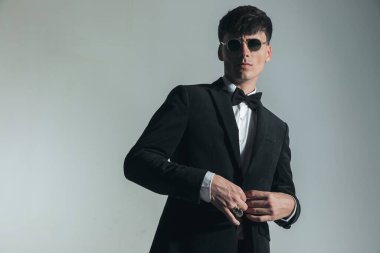 portrait of handsome elegant man with glasses buttoning black tuxedo and posing in a confident way on grey background in studio