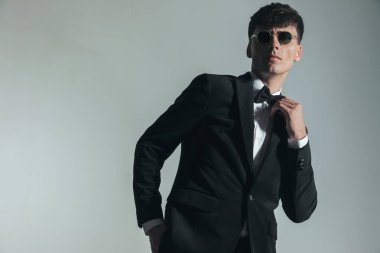 confident elegant groom posing with hand in pocket and fixing bowtie in front of grey background