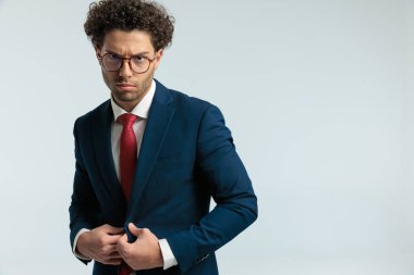 Portrait of elegant businessman opening suit with mad face, standing and wearing eyeglasses against gray studio background