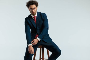 young businessman fixing his sleeve with a mad face, sitting on a chair and wearing eyeglasses against gray studio background