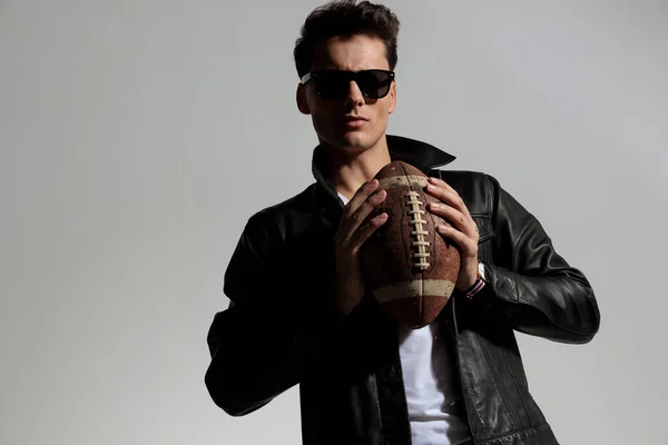 fashion guy in leather jacket playing football and getting ready to throw pigskin ball in front of grey background