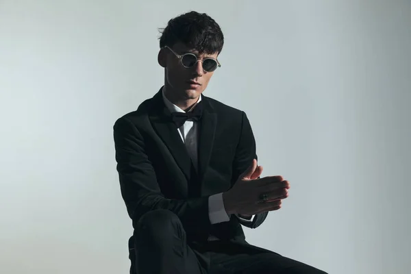 handsome best man with glasses wearing elegant black tuxedo, sitting and rubbing palms while posing in front of grey background