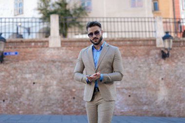 elegant young businessman in suit wearing sunglasses and posing while rubbing palms outdoor